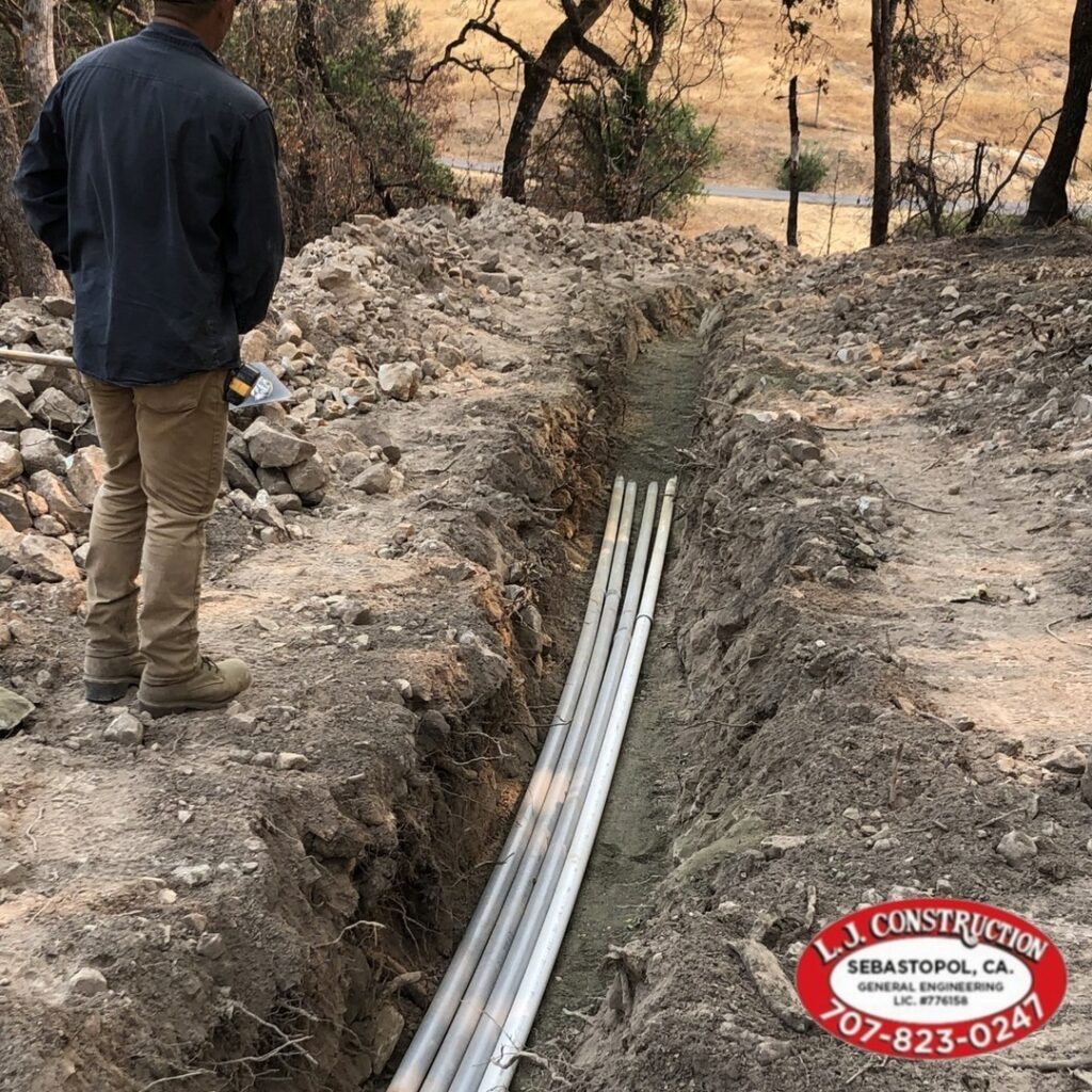 Dirt path with pipes laid out on it, rocks and rubble on either side with a man looking at it.