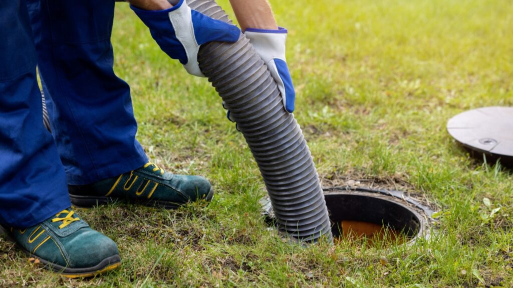 A man in blue protective gloves lowers a large hose into an open septic tank.