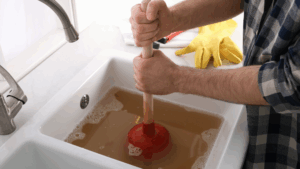 A clogged drain with brown water and a man using a plunger on it.