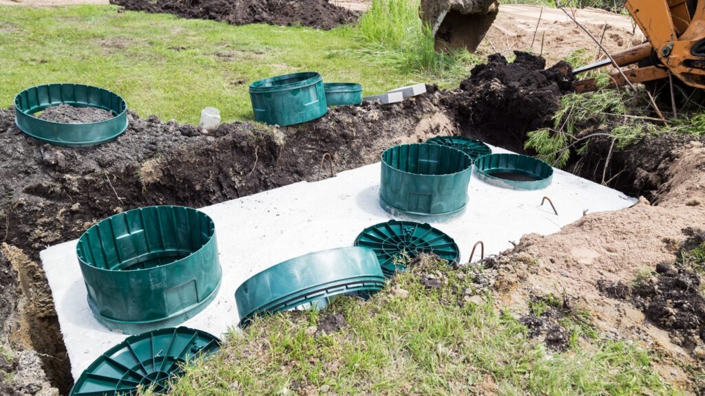 A septic tank in the ground with pipe pieces and an excavator nearby.