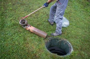 Person in grey pants opening a septic tank and taking out a pipe with a red hook and placing a septic pipe on surrounding grass.