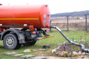 A red septic tank truck connected to a hose, pumping a septic tank in a yard surrounded by rocks and green grass.